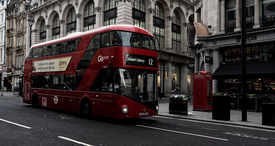 A red double-decker bus on a city street in Dulwich, with large showroom windows reflecting surrounding buildings. The bus displays the route number 12 and the destination Dulwich Library on an electronic sign. Nearby, there's a traditional red telephone booth, black waste bins, and a black lamppost, all positioned on the pavement in front of commercial and residential buildings with elaborate facades. The street turf appears clean, with fresh asphalt and clear road markings, under daylight conditions. This scene illustrates urban transportation and street environment typical of Dulwich, captured in a natural, detailed manner for surface and cityscape depiction. Cleaner Dulwich specializes in domestic and commercial cleaning services to keep such environments hygienic and well-maintained, supporting community cleanliness efforts.