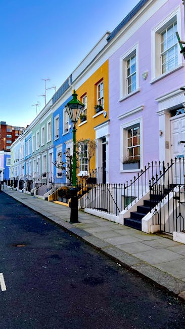 A row of colorful terraced houses on Dulwich Village street, featuring pastel-colored facades in shades of light blue, yellow, and lavender. The houses have white-framed sash windows, black wrought-iron railings along small front steps, and decorative details around doors and windows. A traditional black lamppost stands on the sidewalk, illuminating the street. The scene is bright with clear blue sky, and the pavement appears clean and well-maintained. The image showcases a charming residential area that could benefit from domestic cleaning services like surface cleaning and deep cleaning, which Cleaner Dulwich offers to busy locals in DULWICH.